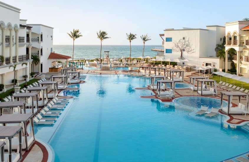 Durante uma tarde ensolarada, área de lazer de um hotel onde ficar em Playa del Carmen com piscinas, espreguiçadeiras, mesas, cadeiras, guarda-sóis, camas e árvores ao redor