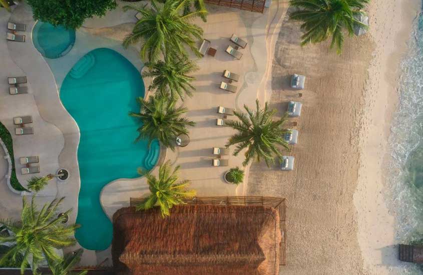 Vista aérea de área de lazer de hotel com piscinas, espreguiçadeiras, árvores e tendas de palha com praia do lado
