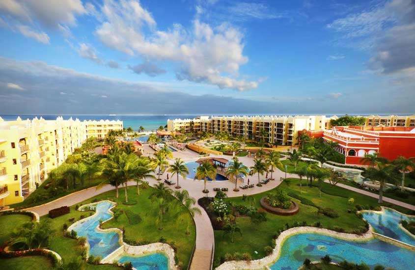 Em um dia de sol, vista aérea de hotel com piscinas, árvores ao redor, partes gramadas e praia na frente