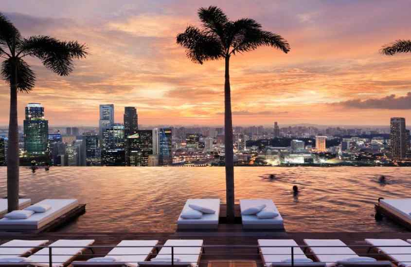 Durante o pôr do sol, área de lazer de um hotel onde ficar em singapura com piscina, camas, árvores e paisagem da cidade na frente