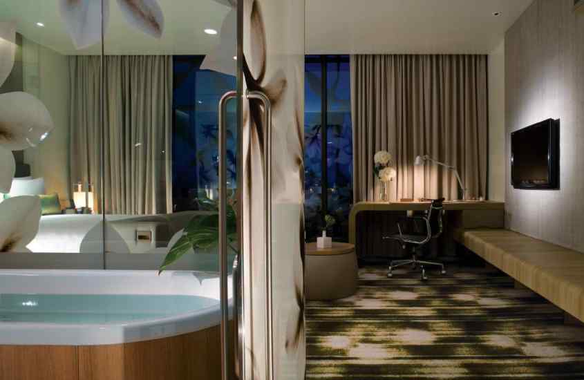 Quarto de um hotel em singapura com banheira, TV, área de trabalho, cama de casal, janela grande acortinada e mesa no meio