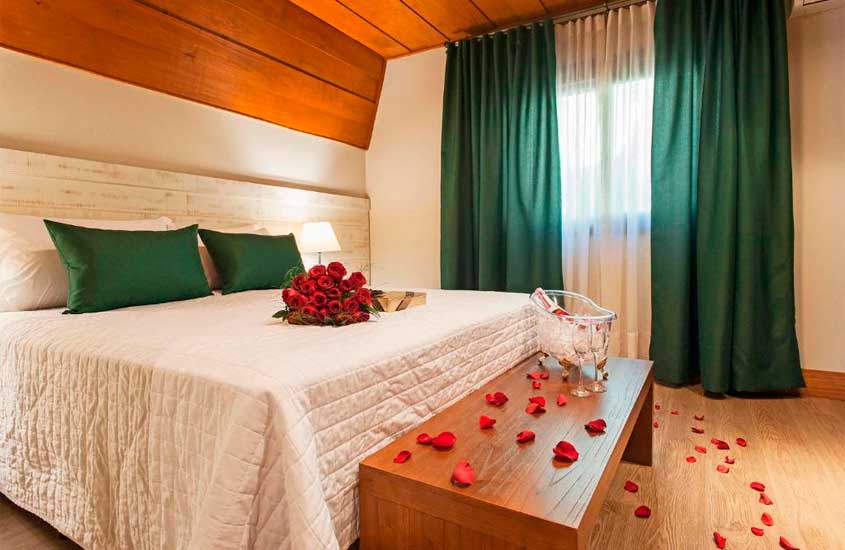 Quarto de hotel com cama de casal, janela grande acortinada, pétalas do rosas espalhadas pelo quarto, balde de champagne, buquê com chocolates na cama