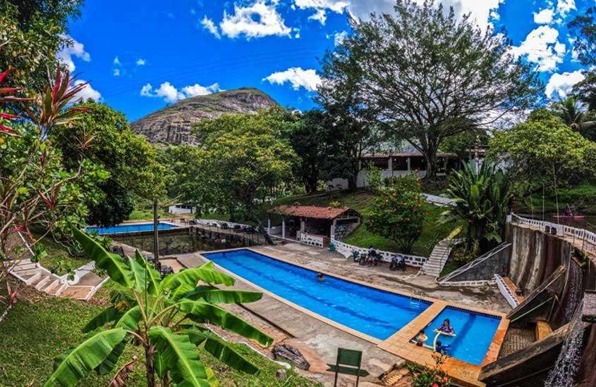 Em um dia de sol, área de lazer de um dos hotéis fazenda em Pernambuco com árvores, plantas e área gramada ao redor, piscinas, montanhas no fundo