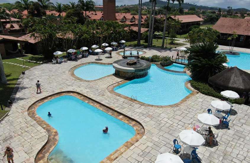 Em um dia de sol, vista aérea de hotel com piscinas, árvores ao redor, mesas, cadeiras e guarda-sóis