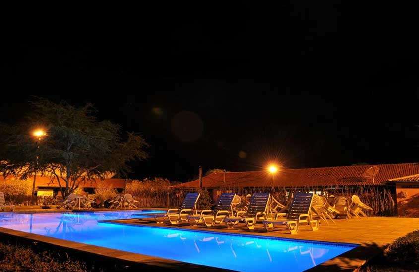 Durante a noite, área de lazer com piscina iluminada, espreguiçadeiras e árvores ao redor