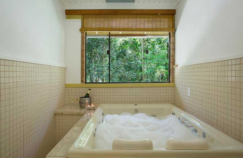 Área de banheira com espuma, janela com vista da natureza, balde de champagne e velas