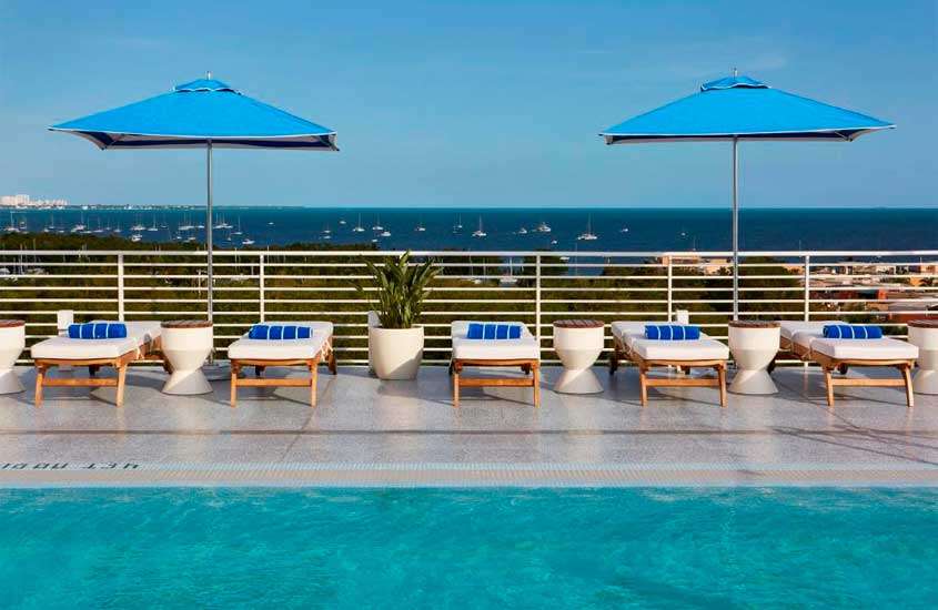 Em um dia de sol, área de lazer de hotel em miami com espreguiçadeiras, guarda-sóis, piscina, planta decorativa e paisagem do mar
