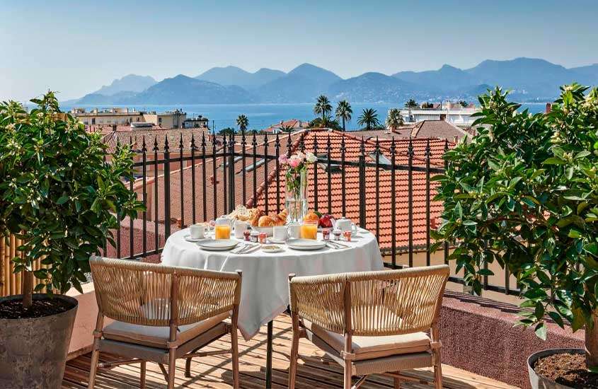Em um dia de sol, cobertura de um hotel onde ficar em Cannes com mesa de café da manhã posta, paisagem do mar e das montanhas e plantas ao redor