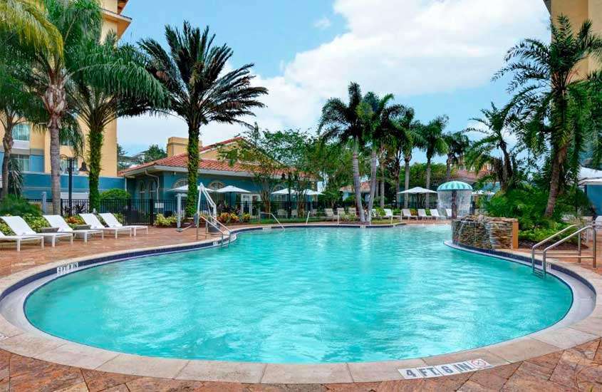 Em um dia de sol, área de lazer de um hotel barato em Orlando perto da disney com piscina, espreguiçadeiras, guarda-sóis, cascata, árvores e plantas ao redor