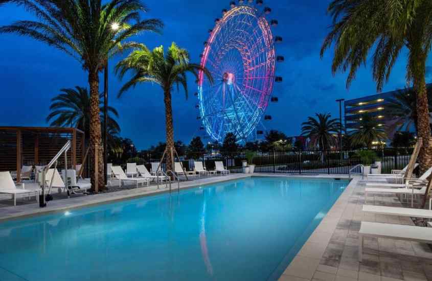 Durante o anoitecer, hotel na international drive com piscina, espreguiçadeiras, árvores e paisagem da roda gigante iluminada