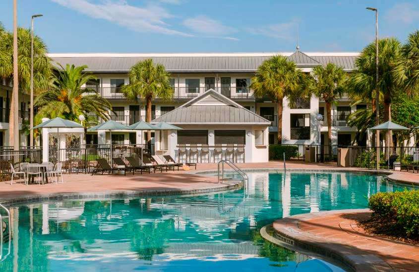 Em um dia de sol, área de lazer de hotel barato em Orlando perto da disney com piscina, espreguiçadeiras, mesas, cadeiras, guarda-sóis e árvores ao redor