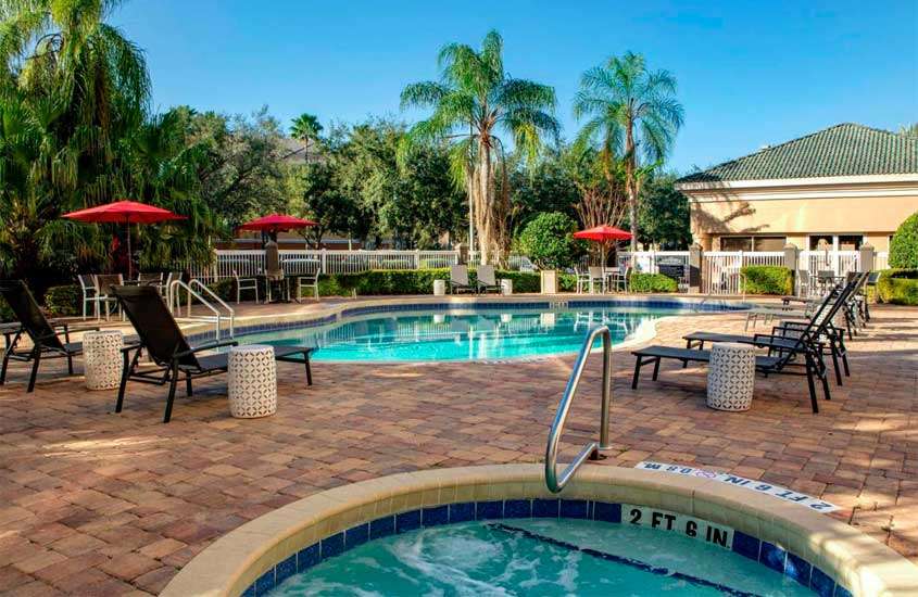 Em um dia de sol, área de lazer de um dos hotéis baratos em Orlando perto da disney com piscina, hidromassagem, espreguiçadeiras, guarda-sóis, plantas e árvores ao redor