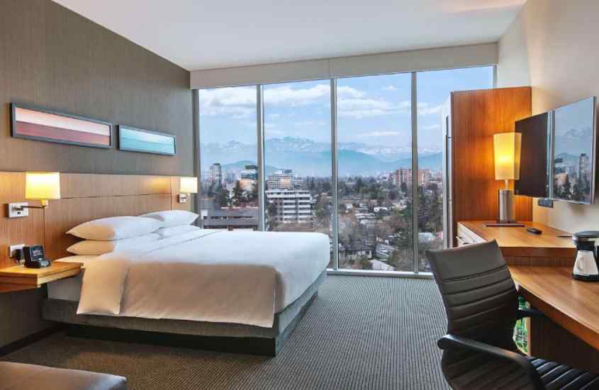 Quarto de um dos hotéis em santiago com vista para a cordilheira, cama de casal, mesa de trabalho, TV, janela grande e quadros decorativos