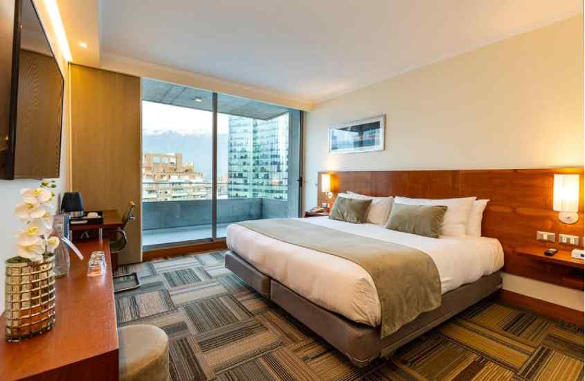 Quarto de hotel com cama de casal, móveis de madeira, quadro decorativo, janela grande acortinada e mesa de trabalho