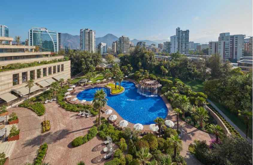Em um dia de sol, vista aérea de hotel em Santiago do Chile com vista para as Cordilheiras, piscina grande, guarda-sóis, árvores e prédios ao redor