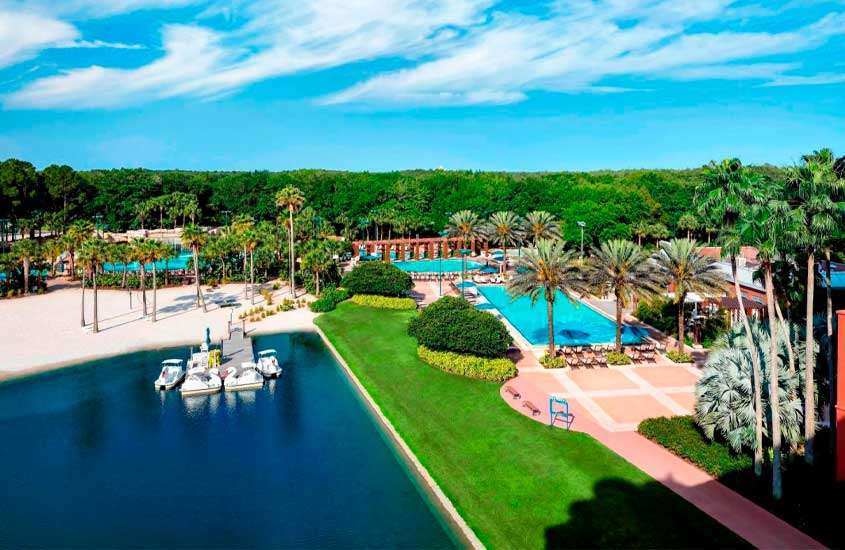 Em um dia de sol, vista aérea de hotel com piscinas, lago com pedalinhos, árvores e plantas ao redor