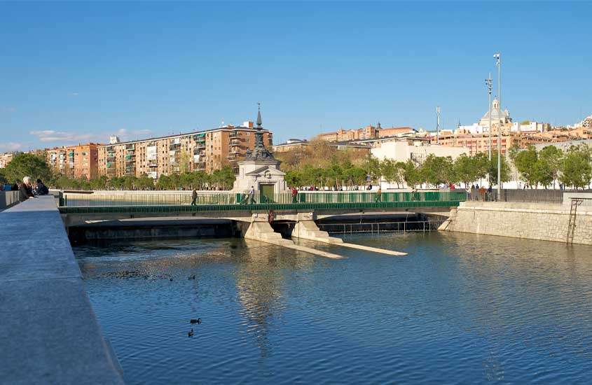Em um dia de sol, ponte com rio embaixo, árvores ao redor e prédios atrás em parque que é uma das opções de o que fazer em Madrid