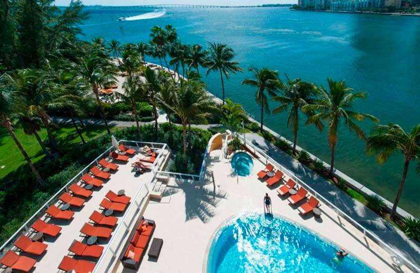 Em um dia de sol, vista aérea da área de lazer de um dos melhores hotéis em Miami com piscinas, espreguiçadeiras, sofás, árvores ao redor, parte gramada e mar do lado