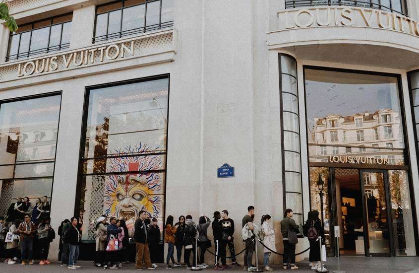 Em um dia de sol, fachada da loja Louis Vuitton com pessoas na fila
