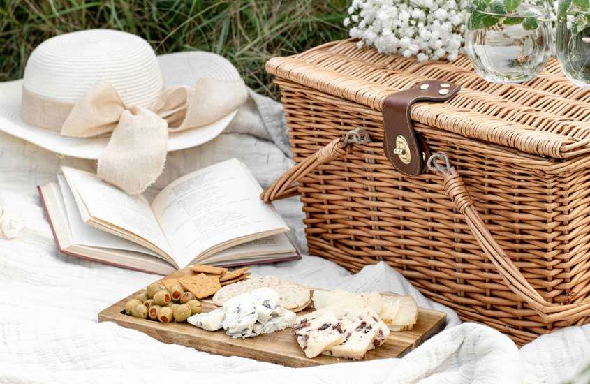 Picnic com queijos, copos de água, flores, cesta, livro e chapéu