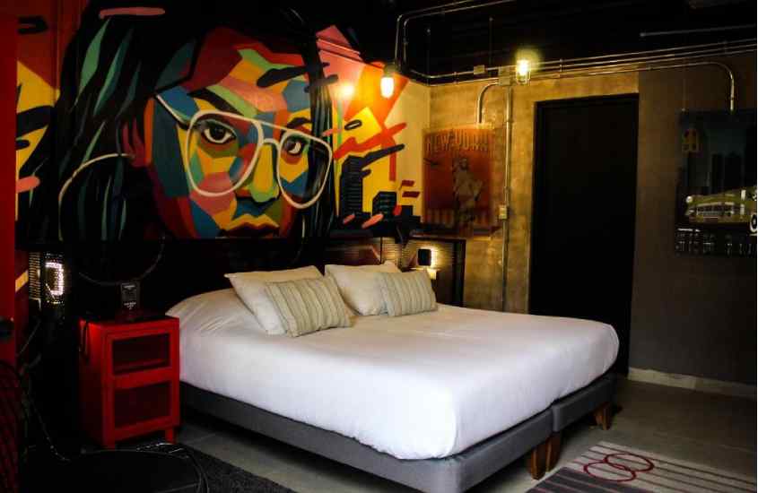 Quarto de hotel com cama de casal, comodas coloridas, parede com pintura, lampadas amarelas e tapete