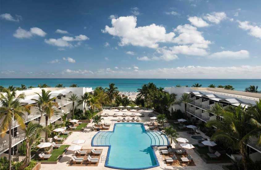 Em um dia de sol, área de lazer de um dos hotéis em Miami Beach com piscina, espreguiçadeiras, guarda-sóis, praia na frente, árvores e parte gramada ao redor