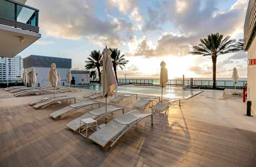 Durante o entardecer, área de lazer de um dos melhores hotéis em Miami Beach com piscina, espreguiçadeiras, árvores ao redor e mar na frente