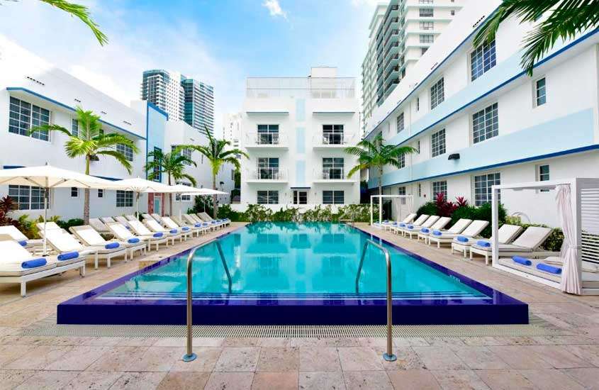 Em uma manhã de sol, área de lazer de hotel em Miami Beach com piscina, espreguiçadeiras, guarda-sóis, cama de descanso, árvores e plantas ao redor