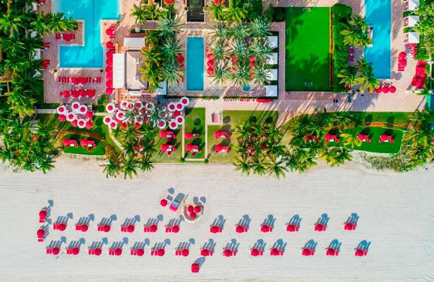 Visão aérea de hotel com guarda-sóis e espreguiçadeiras vermelhas, piscinas, árvores e partes gramada ao redor