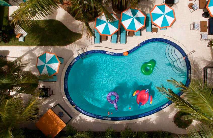 Em um dia de sol, vista aérea da área de lazer do hotel em Miami Beach com piscina, guarda-sóis, espreguiçadeiras, bancos, árvores e parte gramada ao redor
