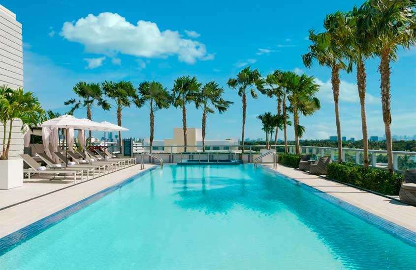Em um dia de sol, área de lazer de hotel em Miami Beach com piscina, espreguiçadeiras, guarda-sóis, árvores e cidade no fundo