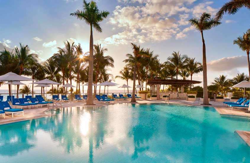 Em um dia de sol, área de lazer de um dos hotéis em Miami Beach com piscina, espreguiçadeiras, guarda-sóis, plantas e árvores ao redor com praia no fundo