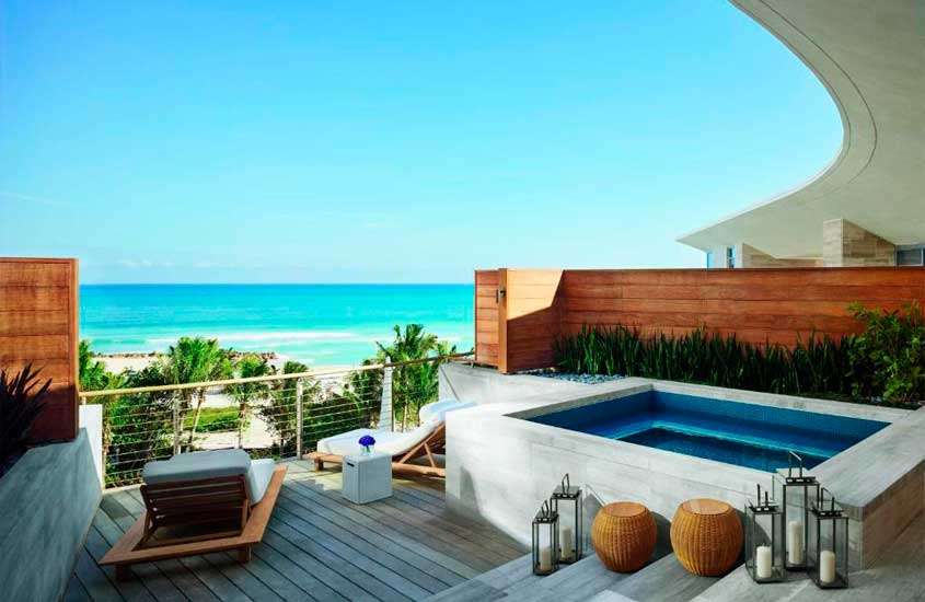 Em um dia de sol, área de lazer de um dos hotéis em Miami Beach com banheira, deck de madeira, espreguiçadeiras, plantas e árvores ao redor e praia na frente