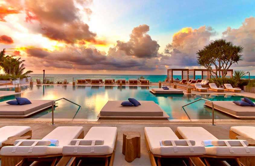 Durante o pôr do sol, área de lazer de um dos hotéis em Miami Beach com piscina grande, camas, espreguiçadeiras, árvores e plantas ao redor e mar na frente