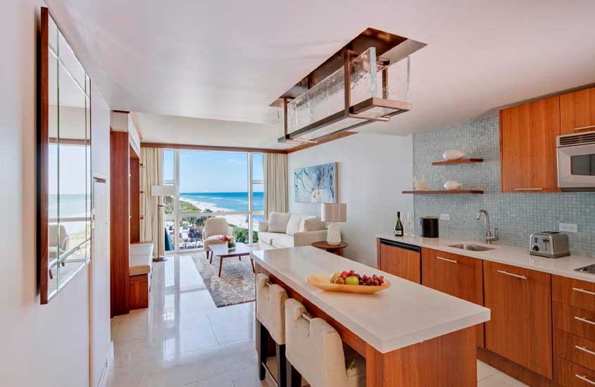 Cozinha com sala conjugada, mesa, cadeiras, poltrona, sofá, tapete, móveis de madeira, espelho, janela grande com paisagem da praia e eletrodomésticos modernos