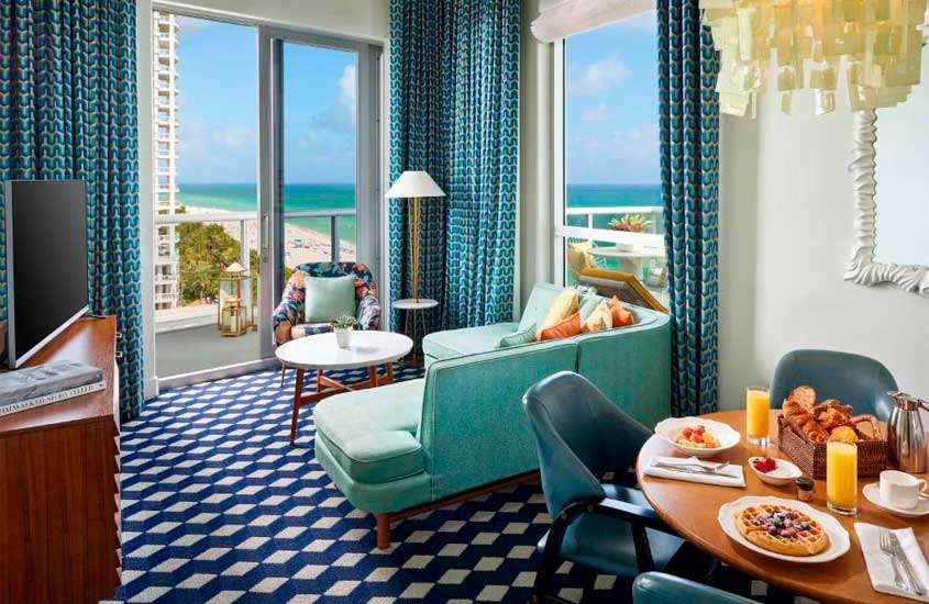 Sala de dois ambientes de um dos hotéis em Miami Beach com mesa de cafe da manha posta, cadeiras, poltrona, sofá, mesa de centro, TV, janela grande acortinada e lustre decorativo