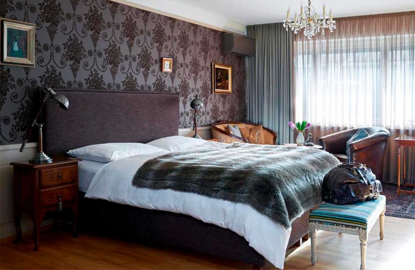 Quarto de hotel com cama de casal, poltronas, ar-condicionado, janela grande acortinada, tapete, cômodas com luminárias e quadros decorativos