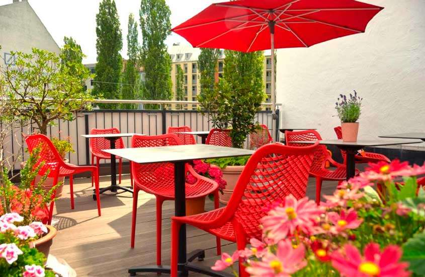 Em um dia de sol, cobertura de um hotel com mesas, cadeiras e guarda-sóis vermelhos, árvores e flores ao redor