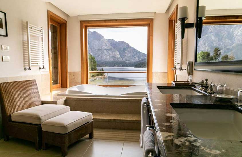 Área de banho de um dos hotéis em Bariloche com poltrona, banheira , janela com paisagem das montanhas, pia e espelho