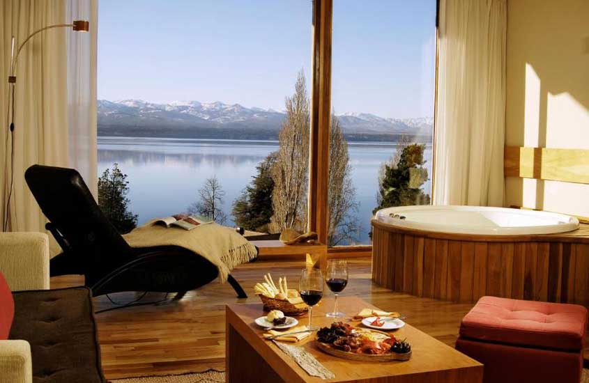 Quarto de um hotel em Bariloche com poltrona, banheira, sofá, puff, mesa posta com petiscos e janela grande com paisagem das montanhas e das árvores