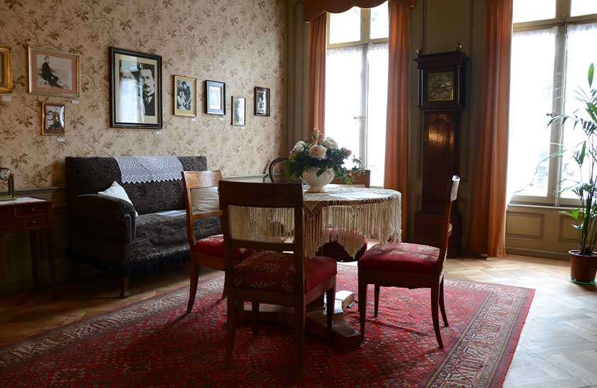 Interior da casa de Einstein com mesa, cadeiras, tapete, sofá, janelas grande, relógio, plantas e quadros decorativos