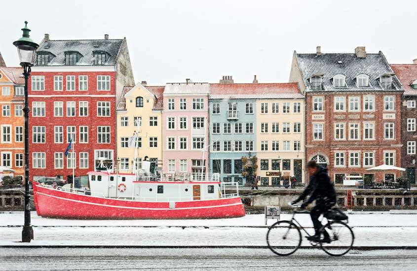 Em um dia de neve, pessoa andando de bicicleta ao lado do canal com prédios coloridos atrás