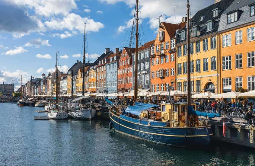 Em um dia de sol com nuvens, porto de Copenhague com barcos, pessoas na margem, guarda-sóis e prédios coloridos ao lado
