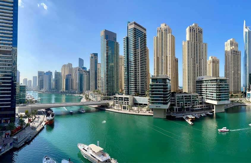 Em um dia de sol, vista aérea da cidade de Dubai com prédios grandes, mar, barcos e árvores ao redor