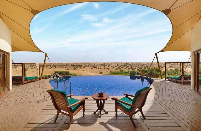 Em um dia ensolarado na cidade de Dubai, hotel com deck de madeira, piscina, cadeiras, mesas, e camas com paisagem do deserto