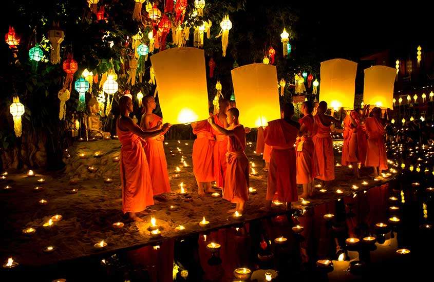 Durante a noite, monges em festival de lanternas com rio na frente, árvore enfeitada e velas ao redor