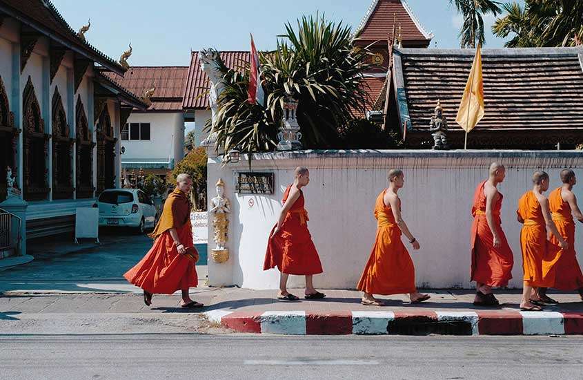 Em um dia de sol, monges de laranja andando pela rua com casas atrás e árvores ao redor