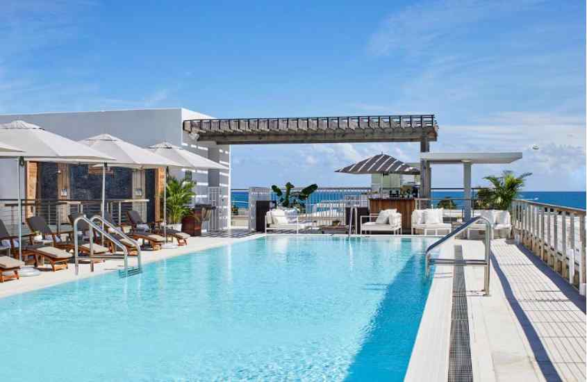 Em um dia de sol, área de lazer de um hotel em Miami Beach em frente à praia com piscina, plantas ao redor, espreguiçadeiras de madeira, guarda-sóis, mesas, cadeiras, bar e paisagem da praia no fundo