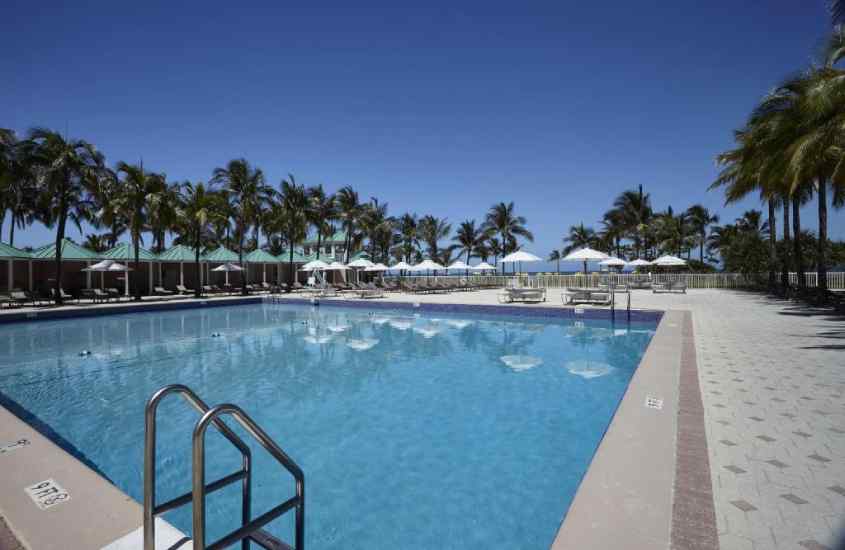 Em um dia de sol, áre de lazer de hotel em Miami Beach em frente à praia, com piscina, espreguiçadeiras, guarda-sóis e árvores ao redor