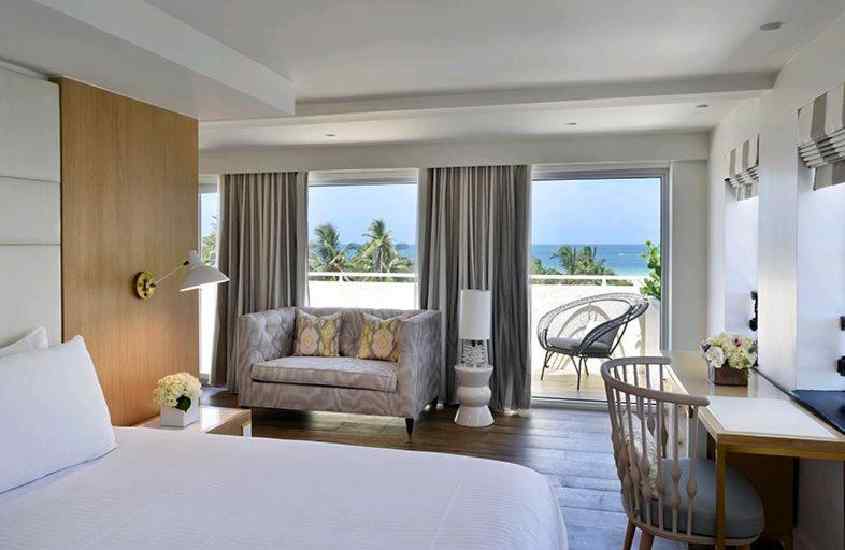 Quarto de hotel em Miami Beach com cama de casal, sofá, cadeiras,flores e luminárias decorativas e varanda